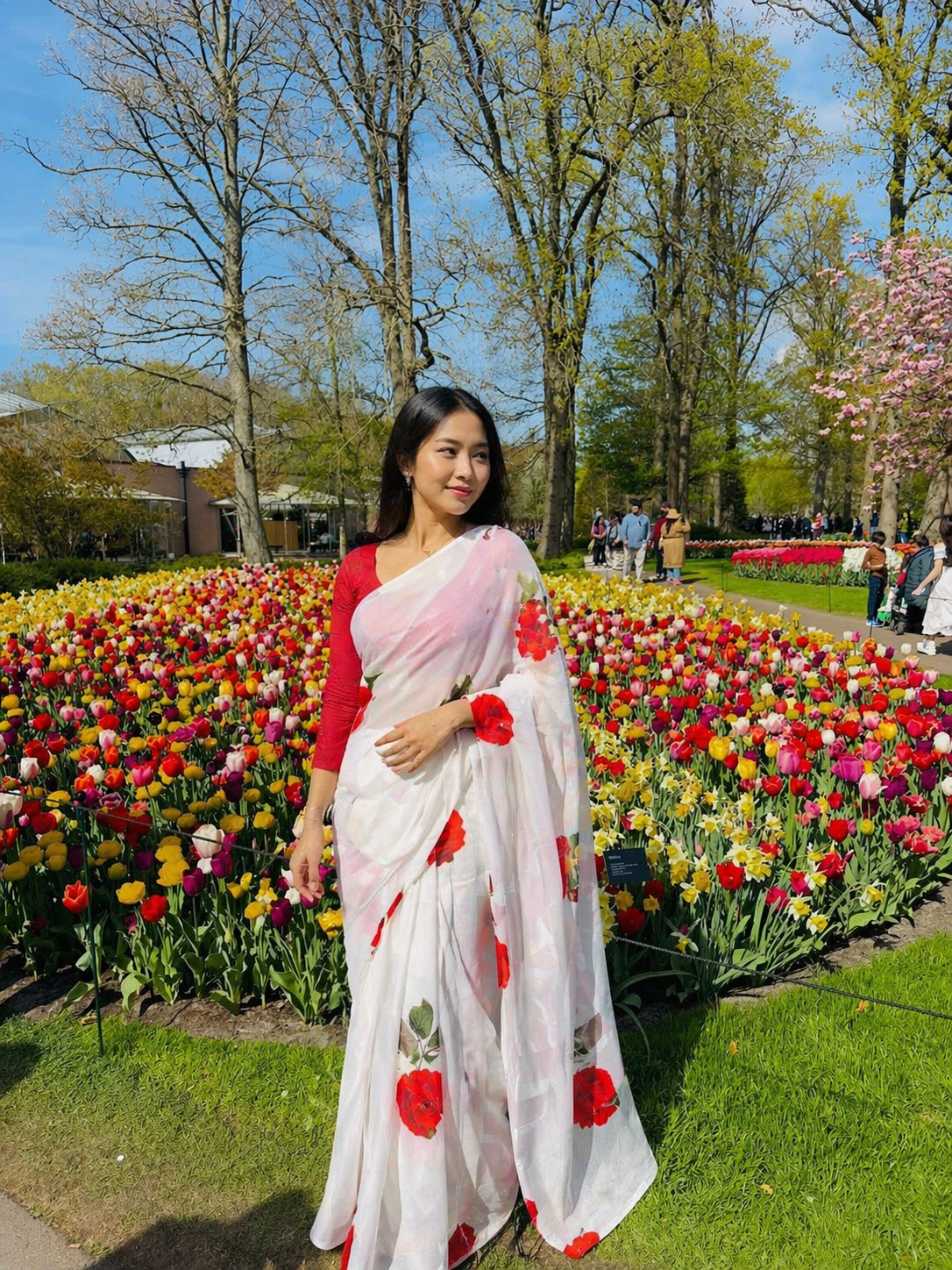 Woman in a white saree with red floral patterns standing in a park with tulips.